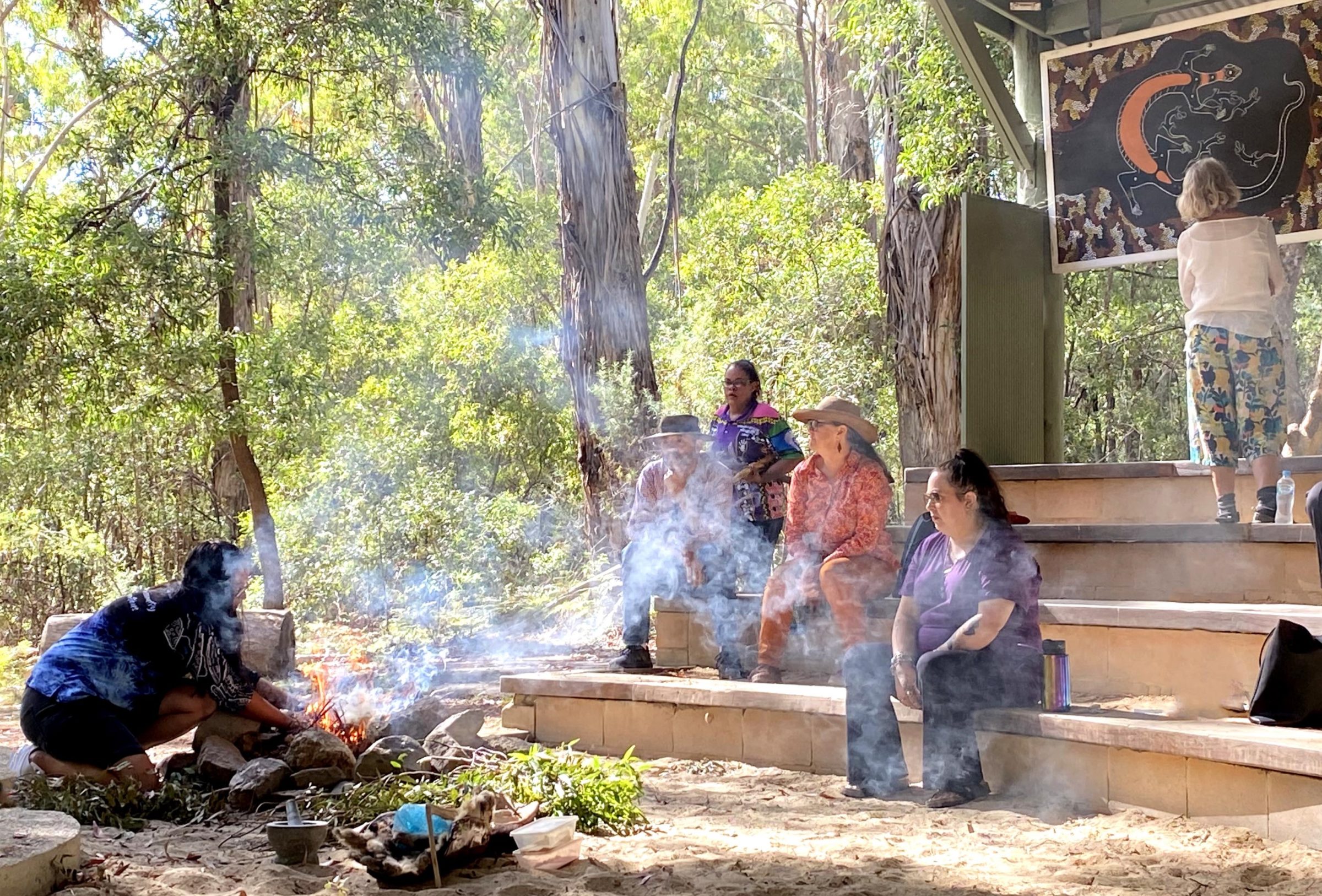 An Aboriginal women stoking a fire for a smoking ceremony while a group of people watch at Tidbinbilla Nature Reserve