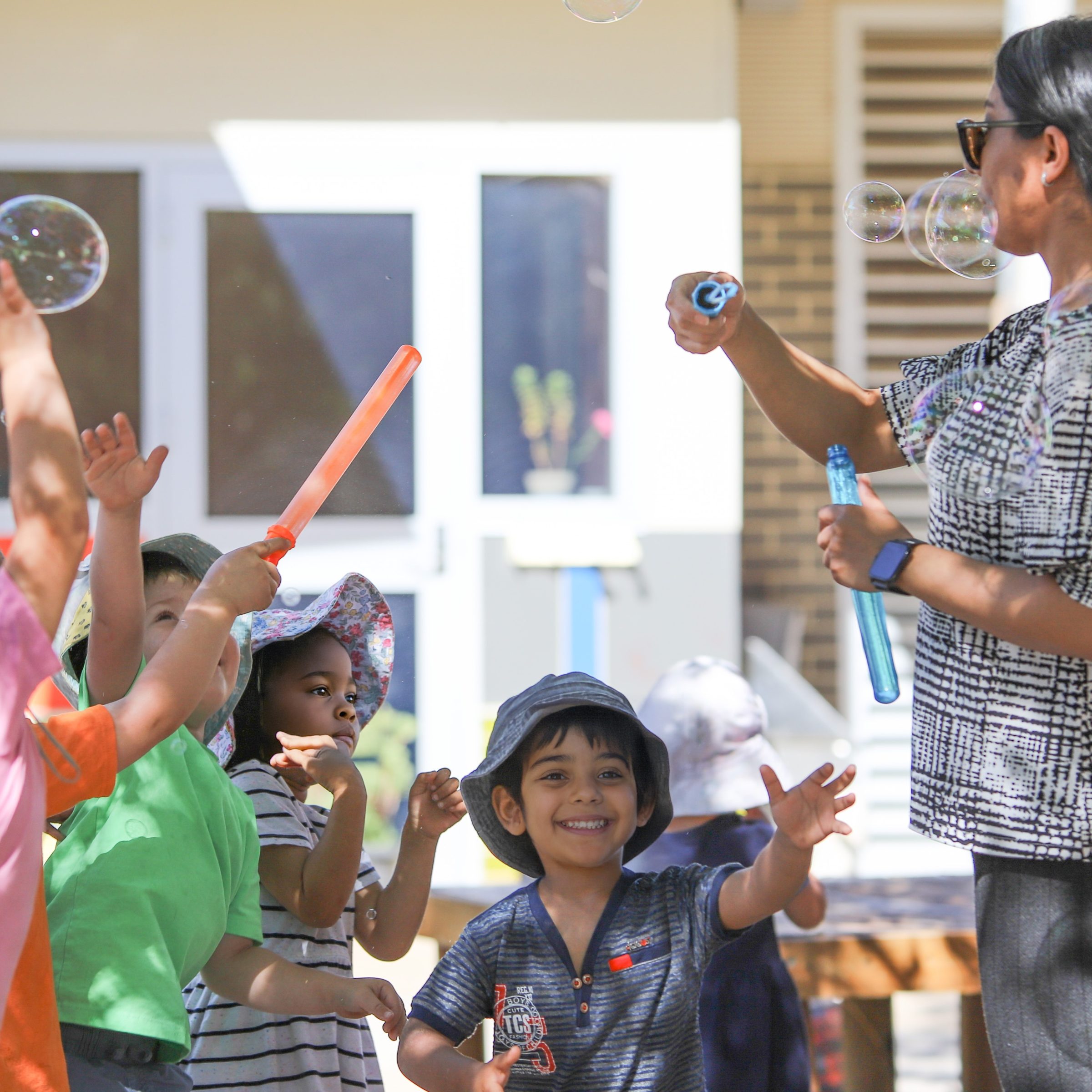 Children happily reaching for bubbles as a teacher blows them toward the group. They are outside and it is sunny at Anglicare's Franklin Early Learning Centre.