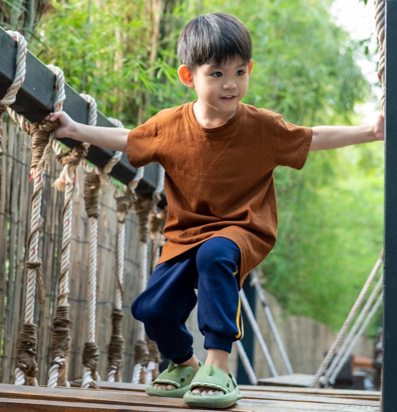 Asian boy carefully getting down from wooden bridge at outdoor playground.